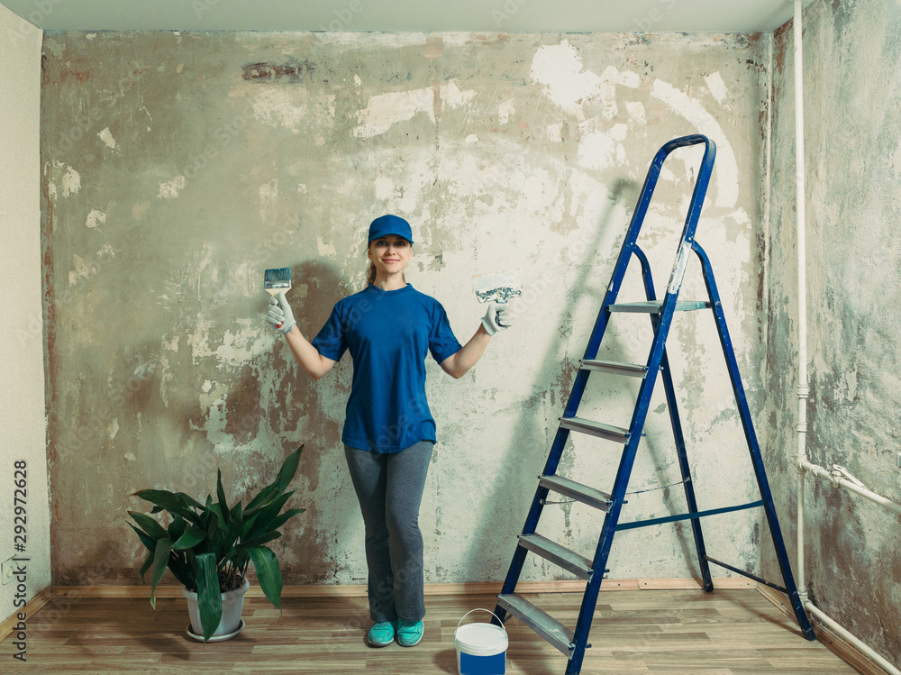 A young woman prepared to do repairs, smiling while standing on the background of a painted wall, in the hands of a trowel and a brush, next to a ladder.