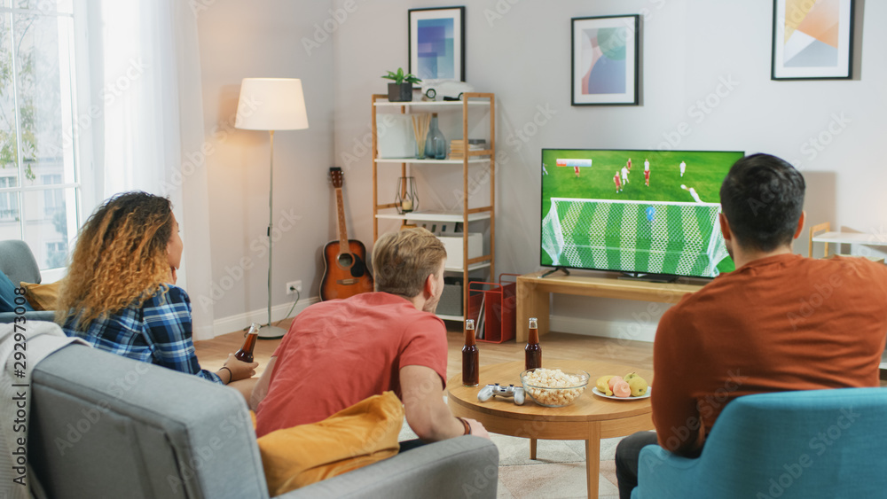Three Sports Fans Sitting on a Couch in the Living Room Watch Important Soccer Match on TV, Cheering For their Team, Waiting for the GOAL that Brings Victory to Their Team. Three Sports Fans Sitting on a Couch in the Living Room Watch Important Soccer Match on TV, Cheering For their Team, Waiting for the GOAL that Brings Victory to Their Team.