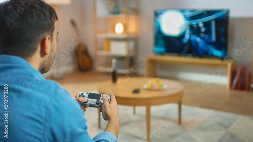 In the Living Room Man Sitting on a Couch Holds Controller Playing in a Console Video Game, 3D Action Shooter Gameplay Shown on TV Screen.