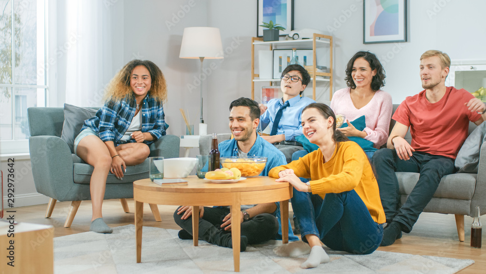 At Home Diverse Group Friends Watching TV Together, Eating Snacks and Drinking Beverage. They Probably Watching Sports Game or Movie. Young People Having Fun Together. At Home Diverse Group Friends Watching TV Together, Eating Snacks and Drinking Beverage. They Probably Watching Sports Game or Movie. Young People Having Fun Together.
