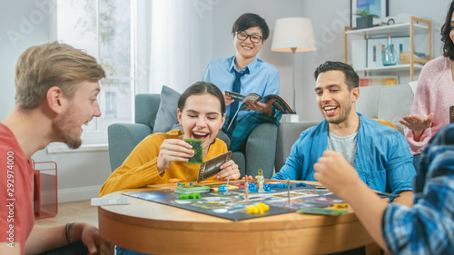 Diverse Group of Guys and Girls Playing in a Strategic Board Game with Cards and Dice. Man Throws Dice Impressively. Cozy Living Room in a Daytime photo