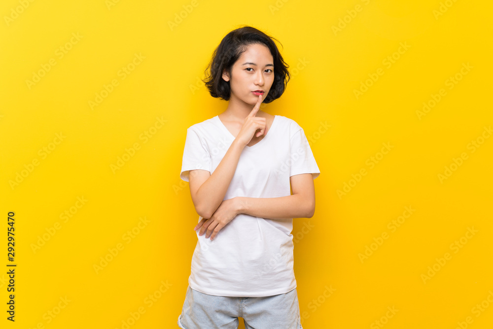 Asian young woman over isolated yellow wall showing a sign of silence gesture putting finger in mouth Asian young woman over isolated yellow wall showing a sign of silence gesture putting finger in mouth