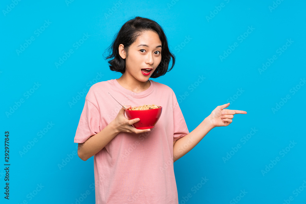 Asian young woman holding a bowl of cereals surprised and pointing finger to the side Asian young woman holding a bowl of cereals surprised and pointing finger to the side