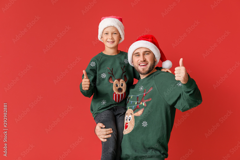 Happy man with his son in Christmas sweaters and Santa hats showing thumb-up on color background Happy man with his son in Christmas sweaters and Santa hats showing thumb-up on color background