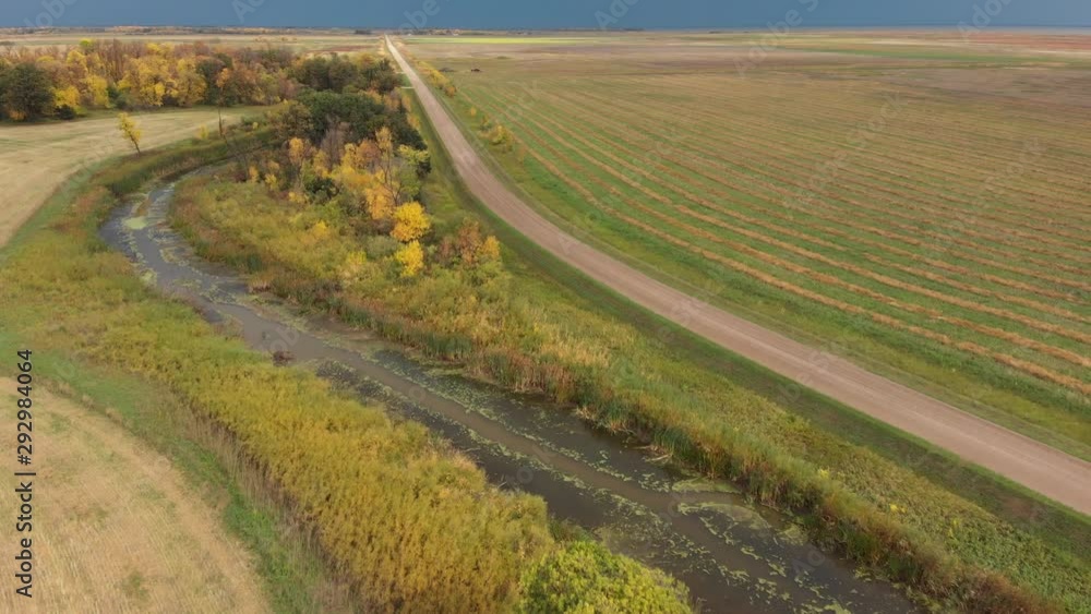 Drone following a gravel road while flying high above farm fields, a river and forests. Dark grey storm clouds are on the horizon. Fall adds beautiful colors to the trees and fields. Drone following a gravel road while flying high above farm fields, a river and forests. Dark grey storm clouds are on the horizon. Fall adds beautiful colors to the trees and fields.