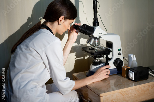 female doctor in white medical uniform with stethoscope on neck looking at specimens under microscope in laboratory photo