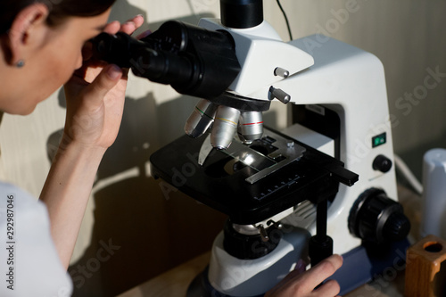 female doctor in white medical uniform with stethoscope on neck looking at specimens under microscope in laboratory photo