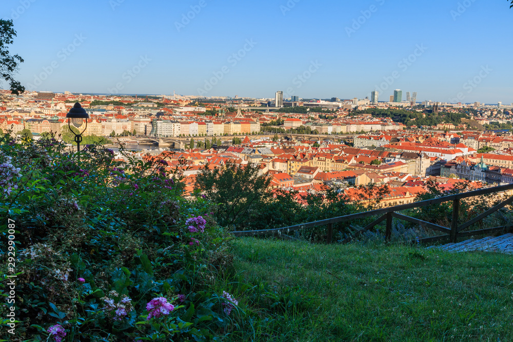 Panoramic view over the rooftops and historical buildings of the district Mala Strana in Prague. In the foreground a green area with purple flowers and street lamp on a sunny day with trees and shrubs