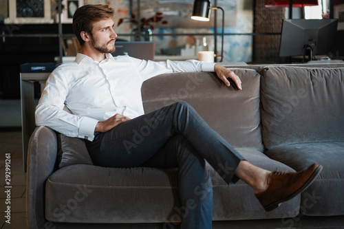 Young and charming businessman is thinking about business while sitting on the sofa in his modern office.