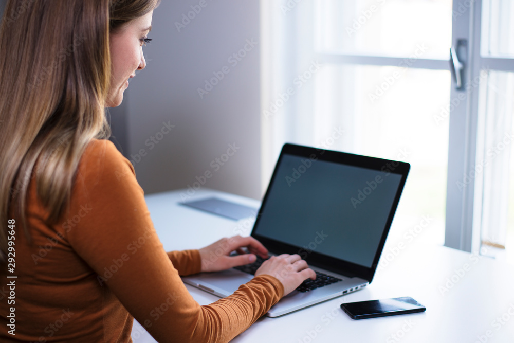 Young female persong typing on laptop on a white desk by the window