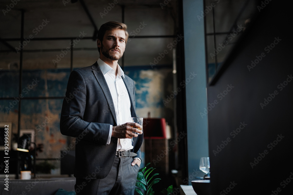 Young handsome businessman stand and hold glass of whiskey in his own office. - obrazy, fototapety, plakaty 