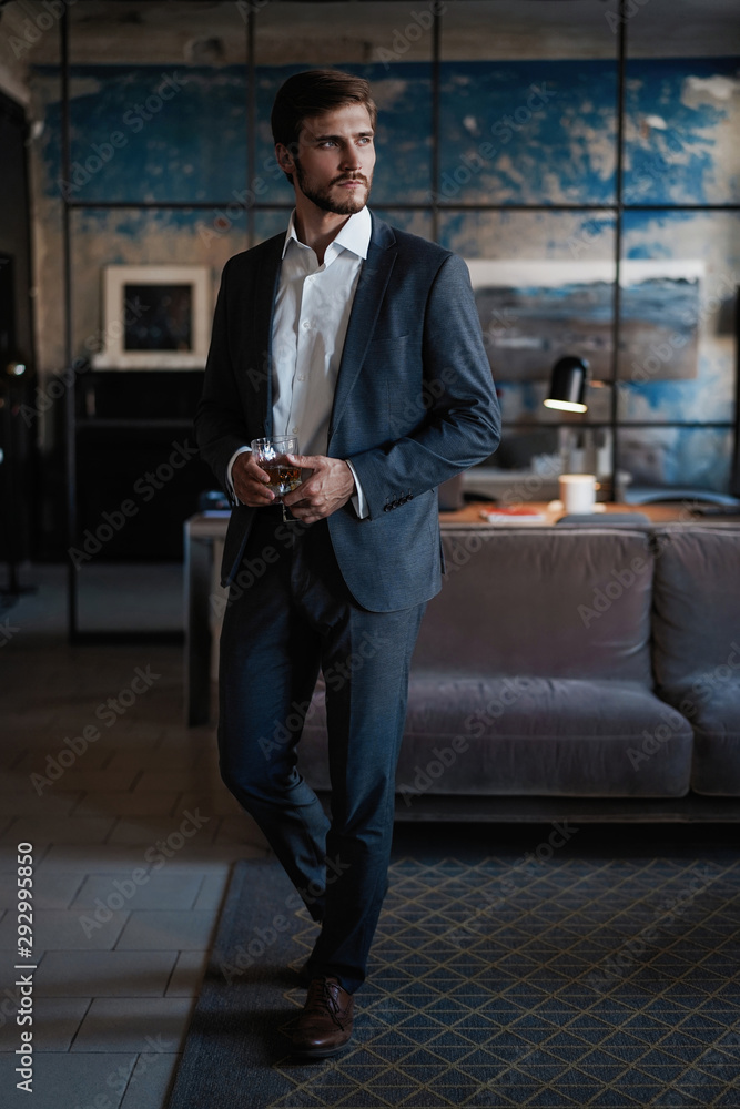 Young handsome businessman stand and hold glass of whiskey in his own office. Young handsome businessman stand and hold glass of whiskey in his own office.