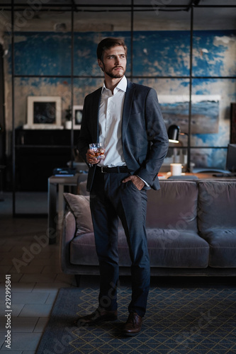 Young handsome businessman stand and hold glass of whiskey in his own office.