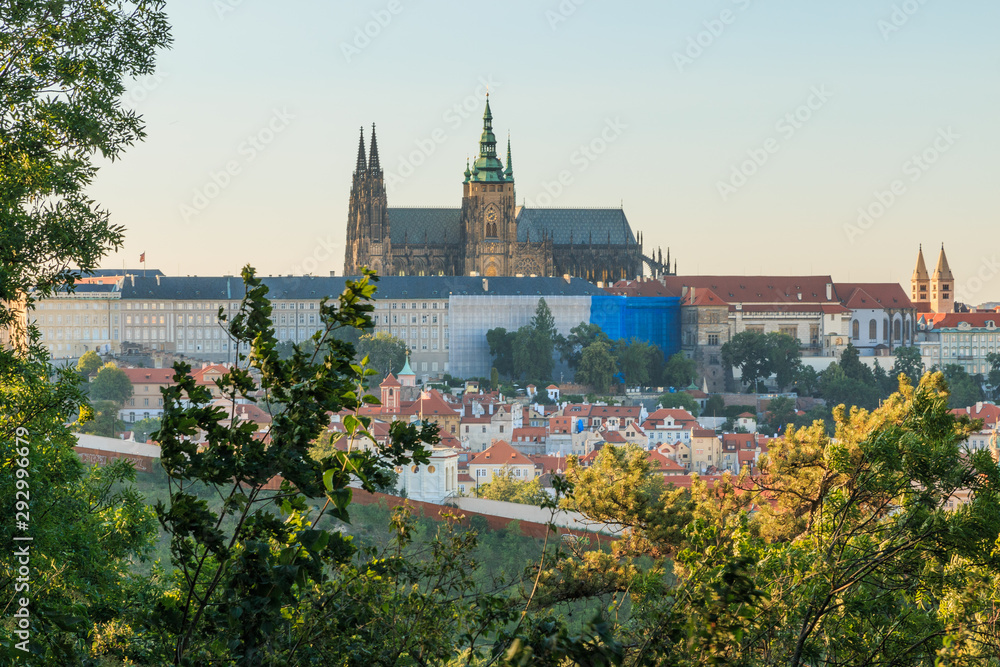 Panoramic view of Prague Castle with the St. Vitus Cathedral above the rooftops of the Mala Strana district from Petrin viewpoint on sunny day with blue sky trees and bushes in the foreground