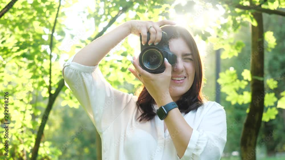 Pretty smiling girl photographer in white shirt is making photos in a park on a soft background of green foliage. A woman photographs model looking at the camera through the rays of the sun at sunset. Pretty smiling girl photographer in white shirt is making photos in a park on a soft background of green foliage. A woman photographs model looking at the camera through the rays of the sun at sunset.