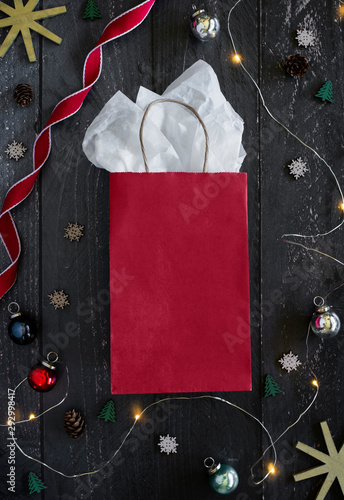 blank red gift bag on dark wooden table surrounded by holiday decor, christmas lights, glass ornaments, snowflakes, pine trees, gold star ornament, red ribbon, gift wrap, copyspace, holiday flat lay
