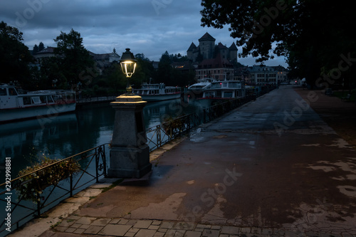 View of the embankment of the Thiou River in Annecy. France. Night time. Street lights are on. The surface of the flowing water is visible. There are old houses and a bridge. Background.\