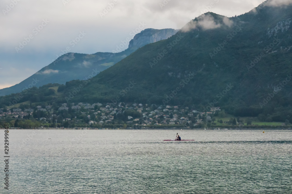 Morning landscape on Lake Annecy, France. In the background are the mountains of the Alps. On the lake in the distance you can see the silhouette of a man kayaking. Morning landscape on Lake Annecy, France. In the background are the mountains of the Alps. On the lake in the distance you can see the silhouette of a man kayaking.