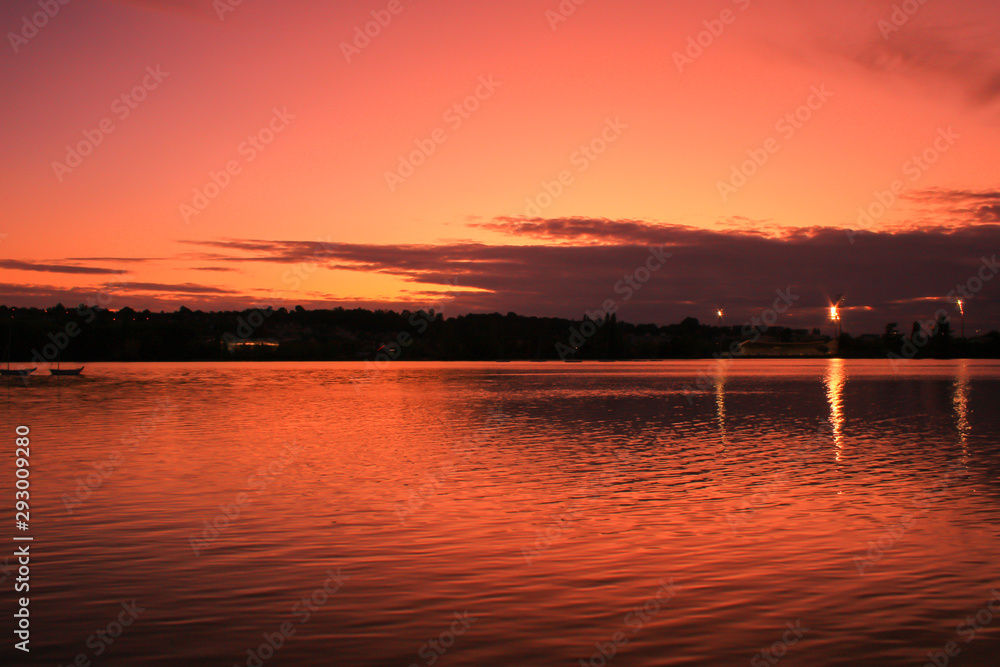 Dark sunset over the water of a lake with symmetry of colorful clouds. Horizon line in background. 