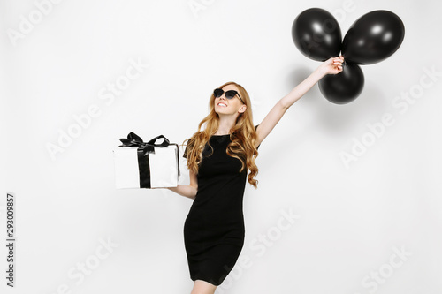 Happy elegant young girl in black dress happily holding gift with black Friday ribbon and black balloons on white background