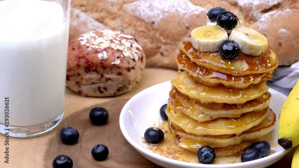 4K wide shot pouring honey or maple syrup on stack of homemade fresh hot pancakes with blueberry and sliced banana on top with a glass of milk for breakfast. Delicious sweet food dessert. 4K wide shot pouring honey or maple syrup on stack of homemade fresh hot pancakes with blueberry and sliced banana on top with a glass of milk for breakfast. Delicious sweet food dessert.