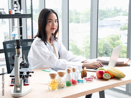 Smiling young asian female nutritionist doctor work on laptop at wooden table with fresh fruit and medicine on foreground in laboratory, healthy concept