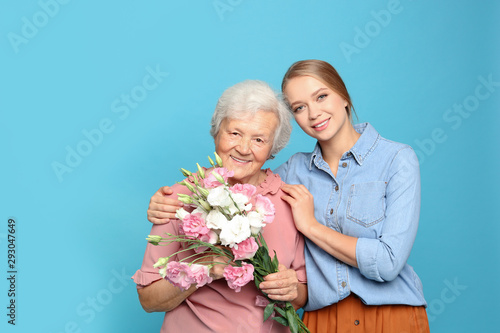 Young woman and her grandmother with flowers on light blue background