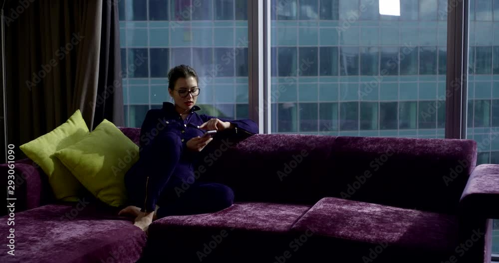 a dark-haired business lady in glasses sits in a beautiful hotel room on a corner purple sofa, against the background of a panoramic window in which skyscrapers are visible. a dark-haired business lady in glasses sits in a beautiful hotel room on a corner purple sofa, against the background of a panoramic window in which skyscrapers are visible.