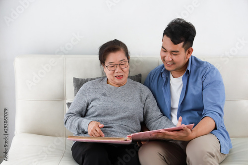 Asian senior woman mother and young man son in blue shirt looking photo album and talking happy smile face  in living room © chayathon2000