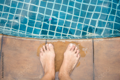 Top view of bare female foot standing beside the edge of blue swimming pool with the petal on the water in sunny day for relaxation