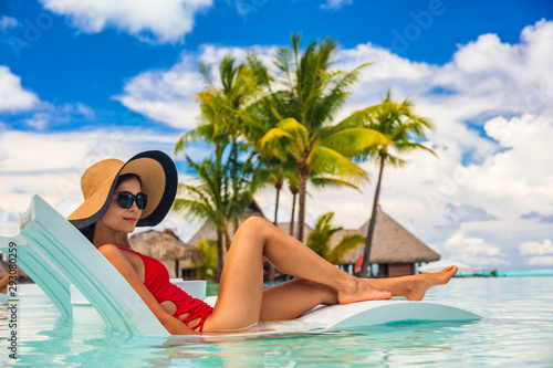 Luxury pool vacation woman sunbathing in lounging chair of high end hotel swimming pool holiday resort for summer getaway. Lady with sun hat and red swimsuit in water lounger.