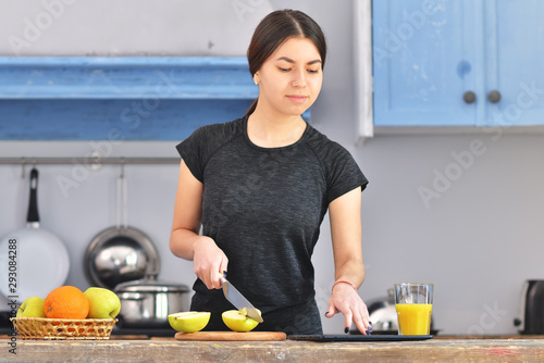 A young girl in a black t-shirt makes a fruit breakfast and cuts apple on a light wooden board in a kitchen. Orange juice and fruit in bowl is standing near. Horizontal photo