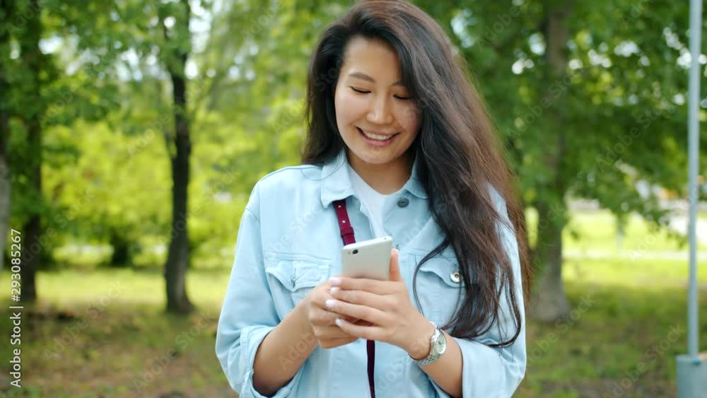 Joyful Asian girl is using smartphone outdoors in city park smiling touching screen standing alone outside. Modern technology, social media and lifestyle concept. Joyful Asian girl is using smartphone outdoors in city park smiling touching screen standing alone outside. Modern technology, social media and lifestyle concept.