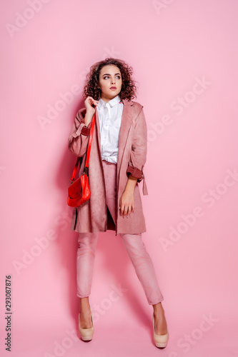 Happy curly brunette girl in coat and bag posing on pink background in studio