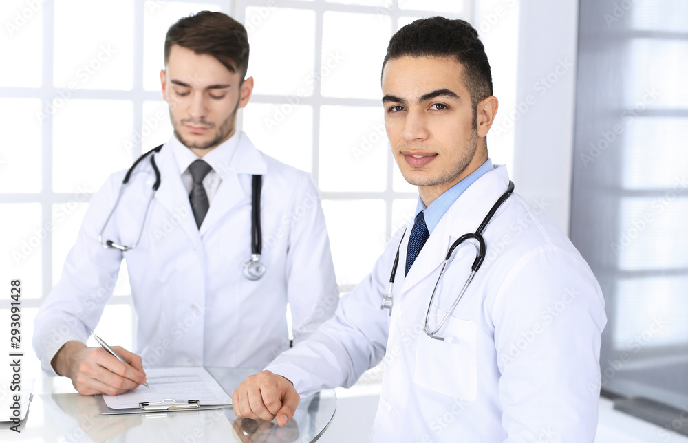 Arab doctor man sitting at the glass desk with caucasian colleague in medical office or clinic. Diverse doctors team, medicine and healthcare concept Arab doctor man sitting at the glass desk with caucasian colleague in medical office or clinic. Diverse doctors team, medicine and healthcare concept