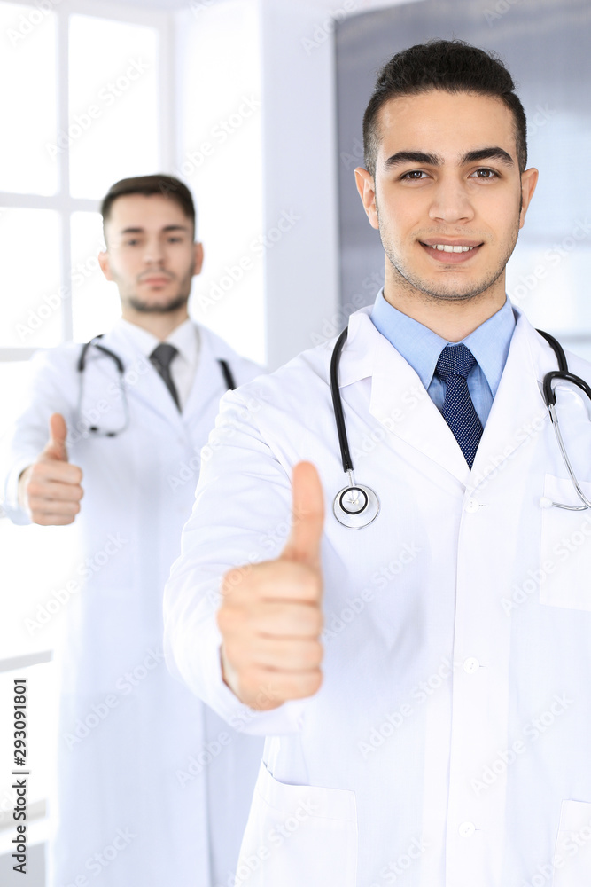 Arab doctor man showing Ok sign with thumbs up with caucasian colleague in medical office or clinic. Diverse doctors team, best treatment, medicine and healthcare concept Arab doctor man showing Ok sign with thumbs up with caucasian colleague in medical office or clinic. Diverse doctors team, best treatment, medicine and healthcare concept