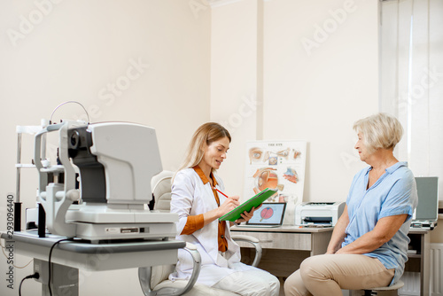 Senior woman patient talking with female ophthalmologist during a medical consultation at the ophthalmologic office. Doctor writing medical history or recipe