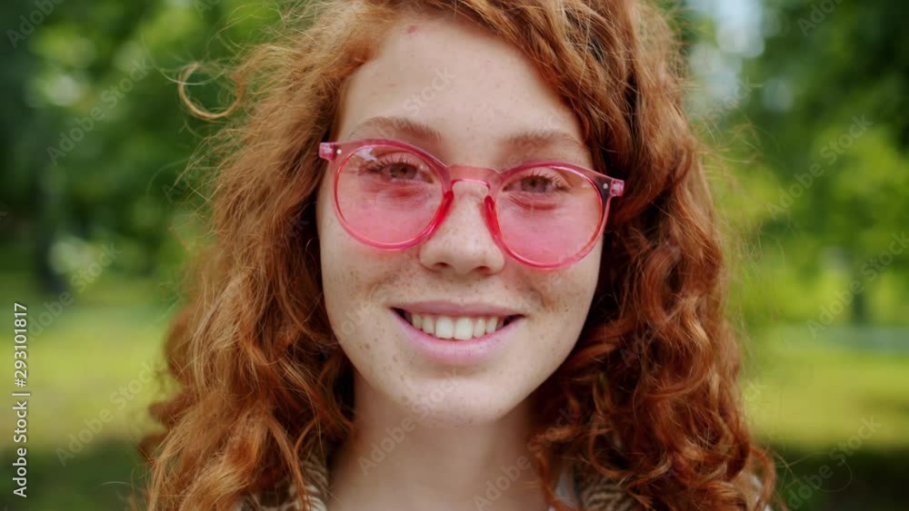 Slow motion of attractive teenager with red curly hair wearing sunglasses smiling in green park alone looking at camera. Youth and positive emotions concept. Slow motion of attractive teenager with red curly hair wearing sunglasses smiling in green park alone looking at camera. Youth and positive emotions concept.