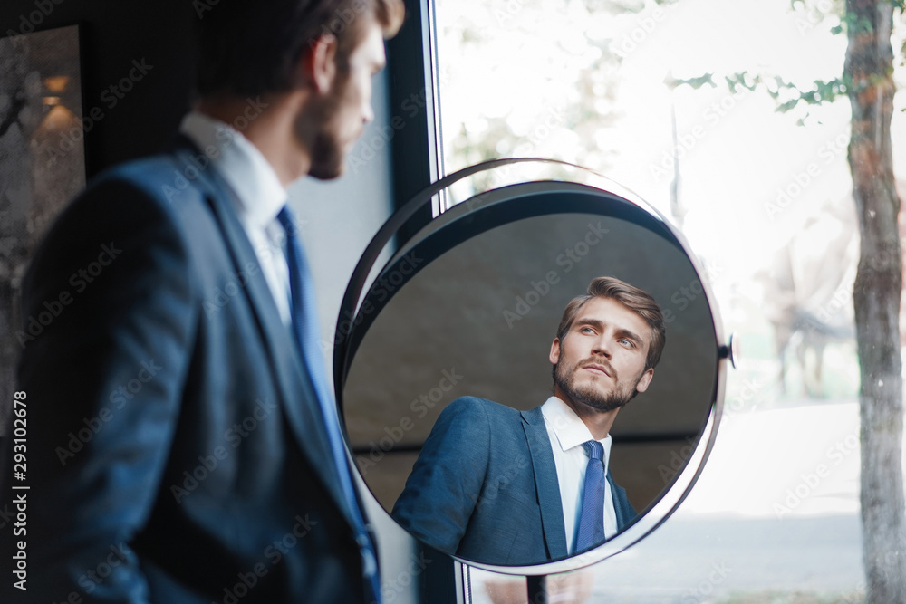 Reflection of handsome young man in full suit standing near the window in front of the mirror indoors - obrazy, fototapety, plakaty 