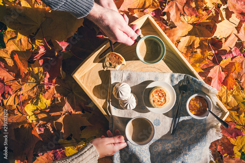 Couple of man and woman having morning breakfast in the autumn garden with colorful maple leaves. cup of coffee, marshmallow jam and a cheese pancake on a wooden tray. Aerial view