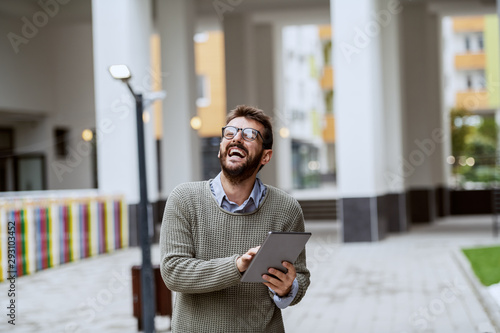 Laughing handsome bearded Caucasian fashionable man in gray sweater and with eyeglasses standing outdoors and using tablet.