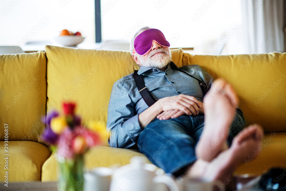 Senior man with eye mask lying on sofa indoors at home, relaxing.