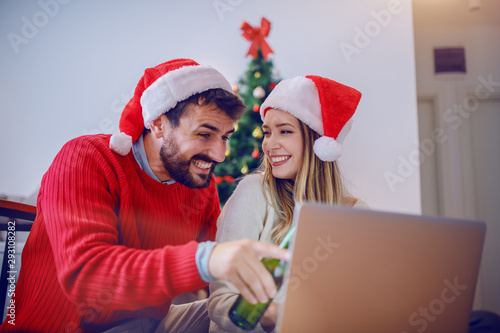 Happy attractive caucasian couple with santa hats on heads and in sweaters sitting on the floor using laptop. Man pointing at laptop while woman looking at man. Christmas holiday concept.