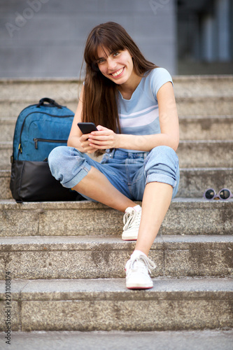 Full body young woman sitting on steps with mobile phone and bag