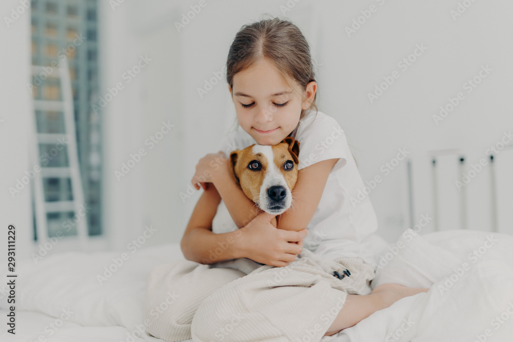 Indoor shot of happy small child embraces favourite pet, dressed in pyjamas, expresses love to her dog, plays with animal after awakening, poses on soft bed. Children and domestic animals concept Indoor shot of happy small child embraces favourite pet, dressed in pyjamas, expresses love to her dog, plays with animal after awakening, poses on soft bed. Children and domestic animals concept