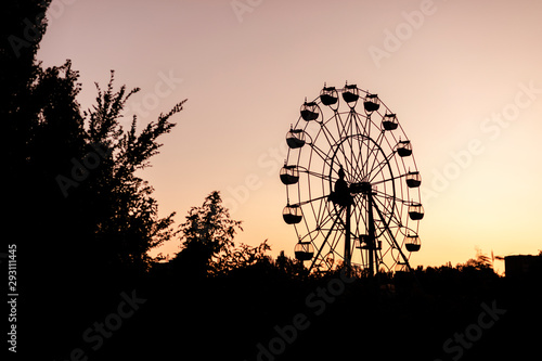 Silhouette of a ferris wheel at sunrise (sunset) of the sun on a background of trees and grass. © COLOR PHOTO