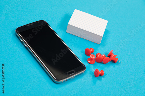 Black mobile phone, a pile of white scratch paper and red push pins are lying on pale-blue surface isolated photo