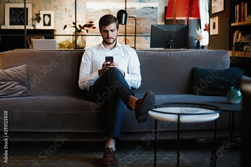 Smiling bearded man making text message on smartphone while sitting sofa at modern office. photo