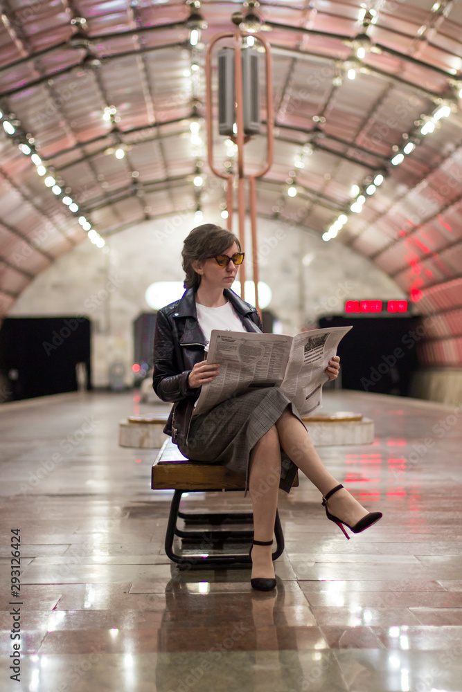 Business woman sitting reading a newspaper. Beautiful girl in a business suit and sunscreen sitting reading a newspaper at the subway station. A woman in a jacket. Success concept - obrazy, fototapety, plakaty 