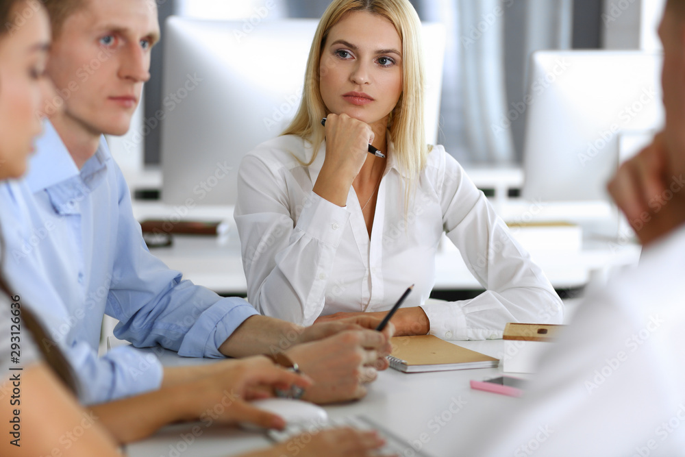 Group of business people discussing questions at meeting. Headshot of blonde businesswoman while smiling to her colleague at office negotiation. Teamwork and cooperation in corporate occupation Group of business people discussing questions at meeting. Headshot of blonde businesswoman while smiling to her colleague at office negotiation. Teamwork and cooperation in corporate occupation